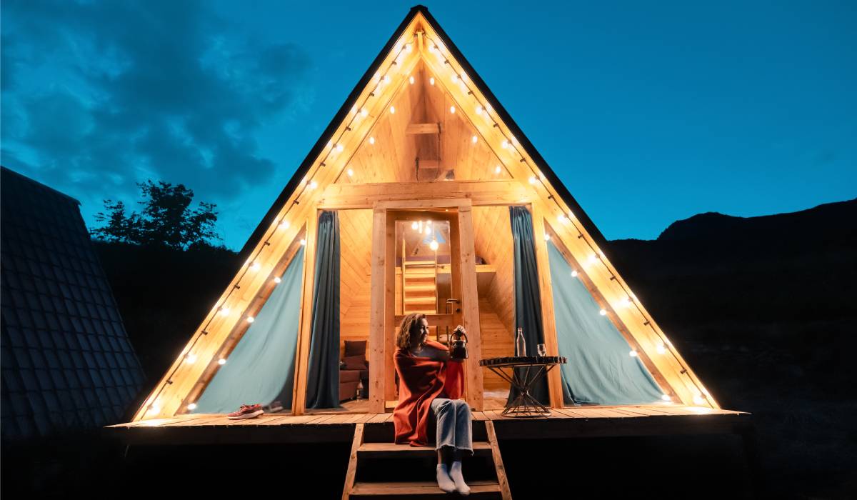 A woman sitting on the porch of an outdoor glamping tent. The tent is lit up and the night sky is slightly cloudy.