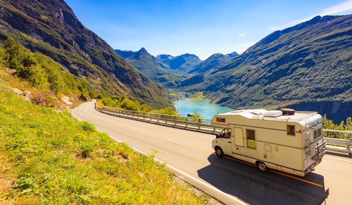 An RV traveling across a two-lane mountainous highway. There's a small lake, a valley, and the sun is shining.