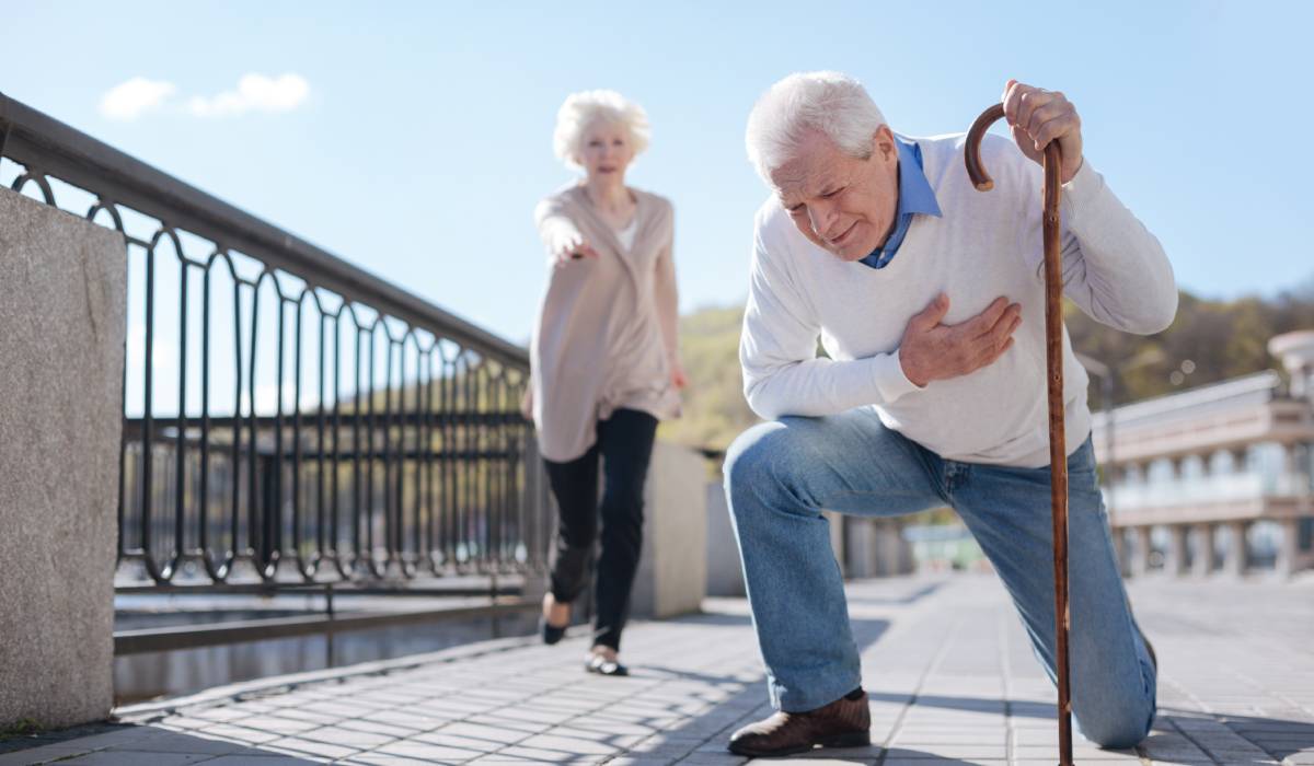 An elderly man with a cane crouches down outside while clutching his chest. An elderly woman is behind him.