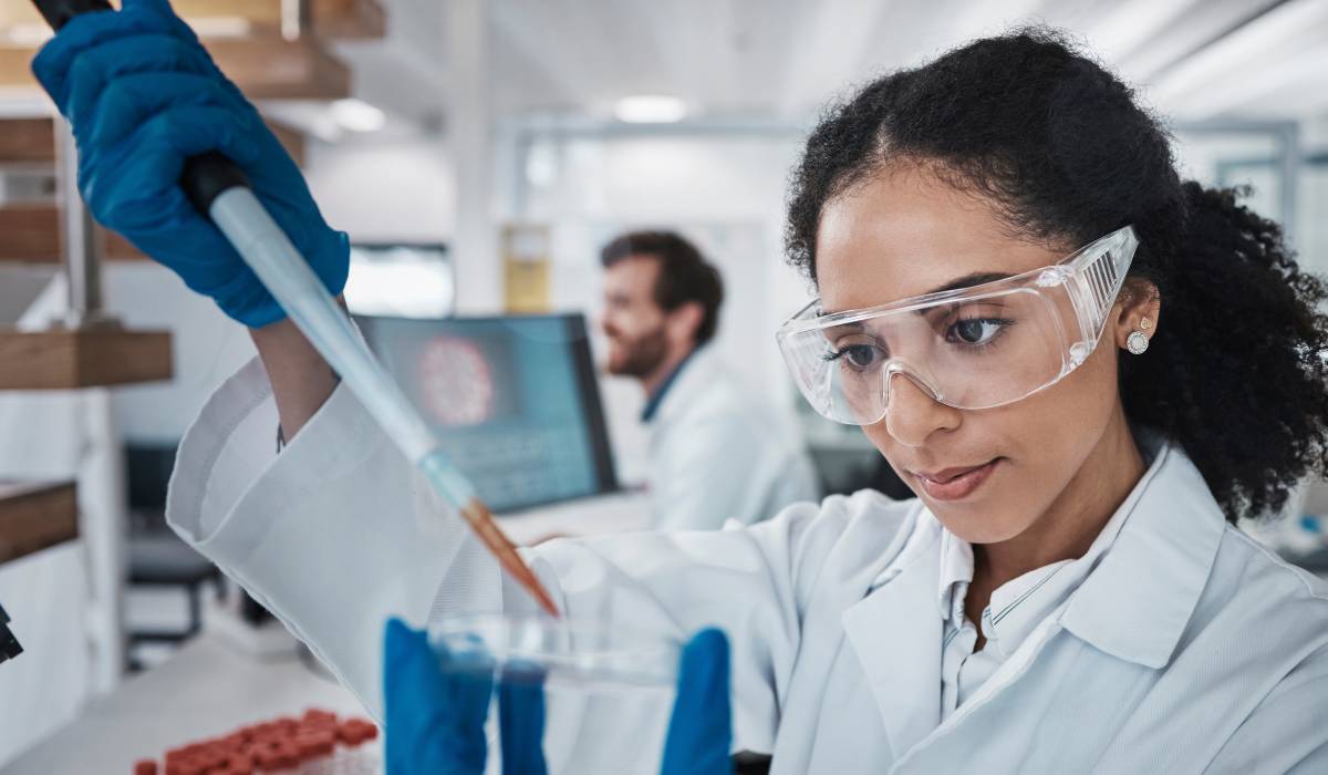 Female scientist wearing white robe, safety goggles, and blue latex gloves, holding a pipette over a petri dish.