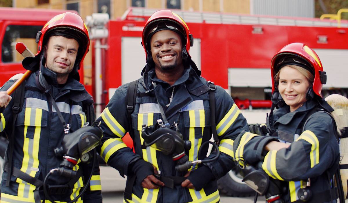 Three firefighters dressed in black protective clothing and red helmets stand in front of a fire truck and smile.